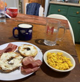 A cup of coffee, a bowl of scrambled eggs, a bagel with cream cheese, turkey bacon, and a mug of water on a table