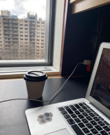 A to-go cup of coffee and an open laptop in front of a window at the library.