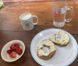 Cut up strawberries, a bagel and cream cheese, a cup of coffee, and a mug of water on a kitchen table