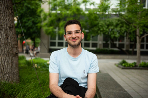 A student seated outside on a bench on campus on summer day