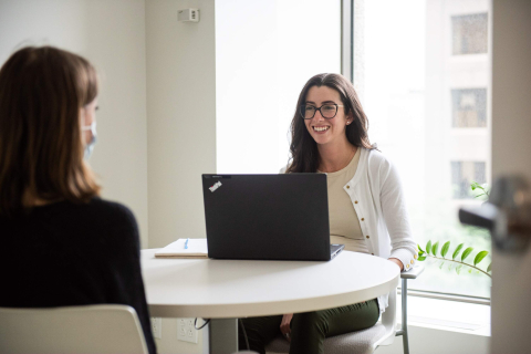 An advisor and student meet at a round office table in front of a window on a bright day