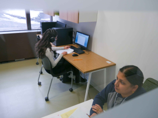 One student wearing earmuffs and one invigilator seated in a small exam room.