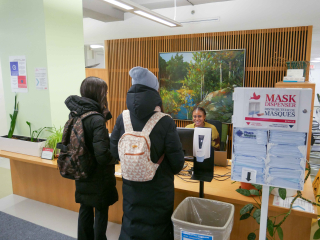 Two students are greeted by a staff member at the reception desk of the Exam Centre. The reception desk is low with overhanging countertop for ease of access.