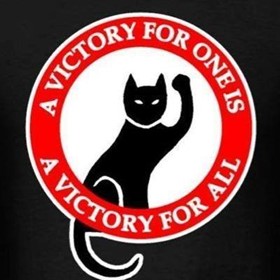 Image of a black cat on black background, surrounded by a red circle and the words "A victory for one is a victory for all" . The cat is raising its left arm in a fist.