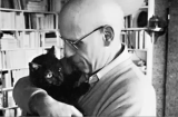 Bald and bespectacled French theorist Michel Foucault cuddles a black cat in a room with full bookshelves