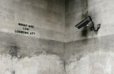 black and white photo of a building corner, surveillance camera mounted on the right pointed at the words "What are you loking at?" printed at the oppopsite wall