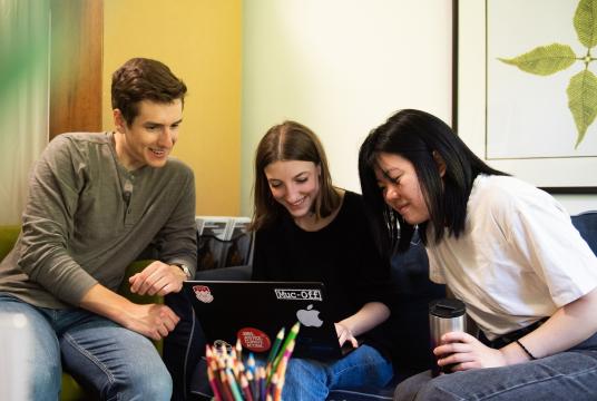 Three students sit together working on a laptop and smiling