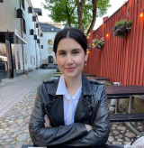 smiling student arms crossed leather black jacket on white blouse cobbled stone path with red fence to the right with hanging baskets and light bulbs