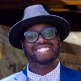 A picture containing a person with glasses and a hat smiling