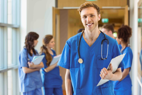 A medical resident in blue scrubs with stethoscope around neck holding a clipboard