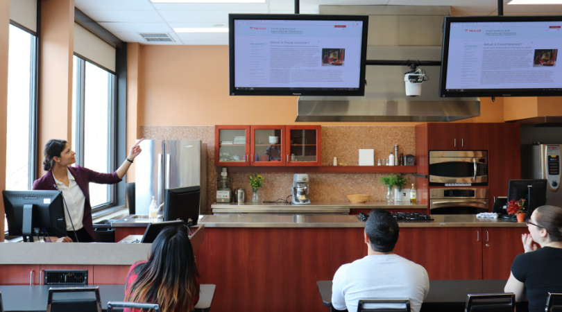 A presenter points at a screen in front of attentive participants. In the background, we see the kitchen of the demonstration room. A camera is positioned above the counter.