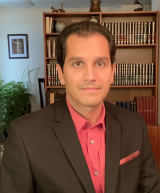 Dr. Labos in red shirt and dark jacket sits in front of a full book shelf