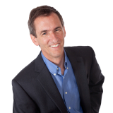 Smiling dark-haired man in a blue suit, no tie, against a white background.