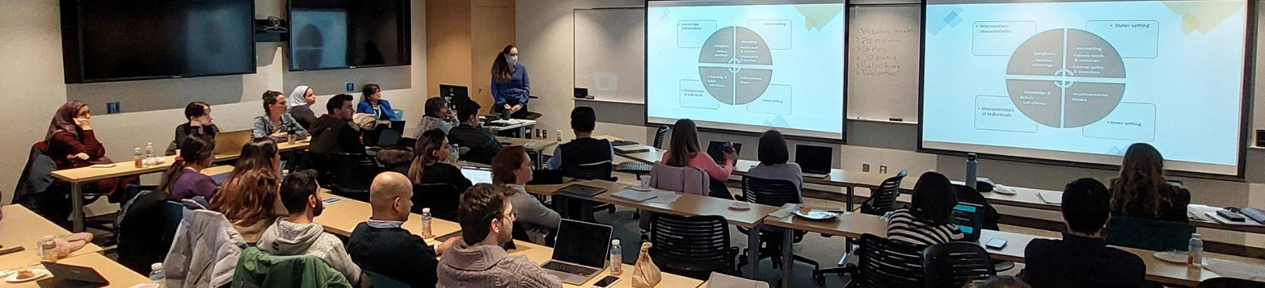 Photo taken from the back of a lecture room during a presentation: people are sitting down looking at two screen projectors while someone is presenting