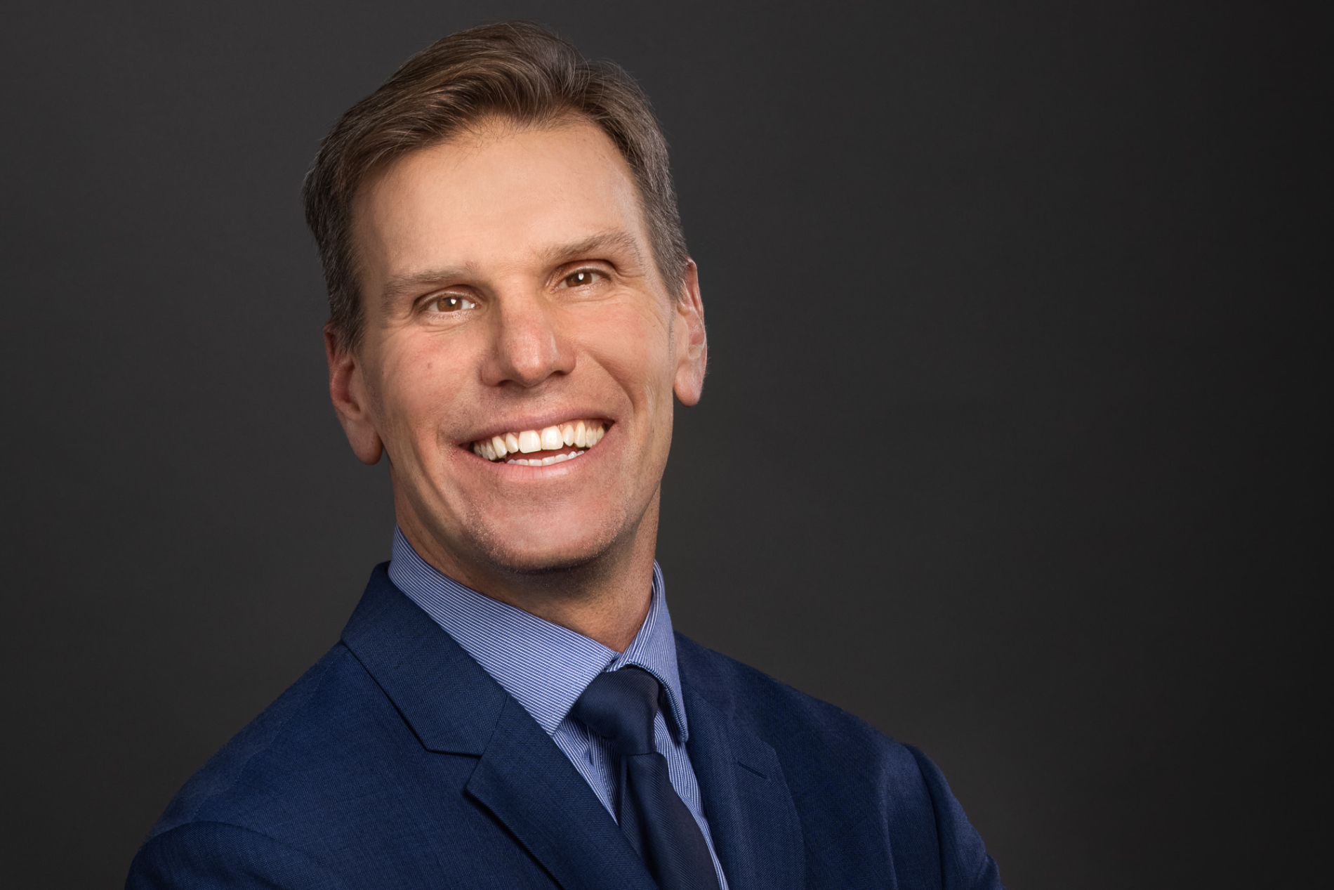 A blond-haired man in a blue shirt and navy jacket and tie smiles widely.
