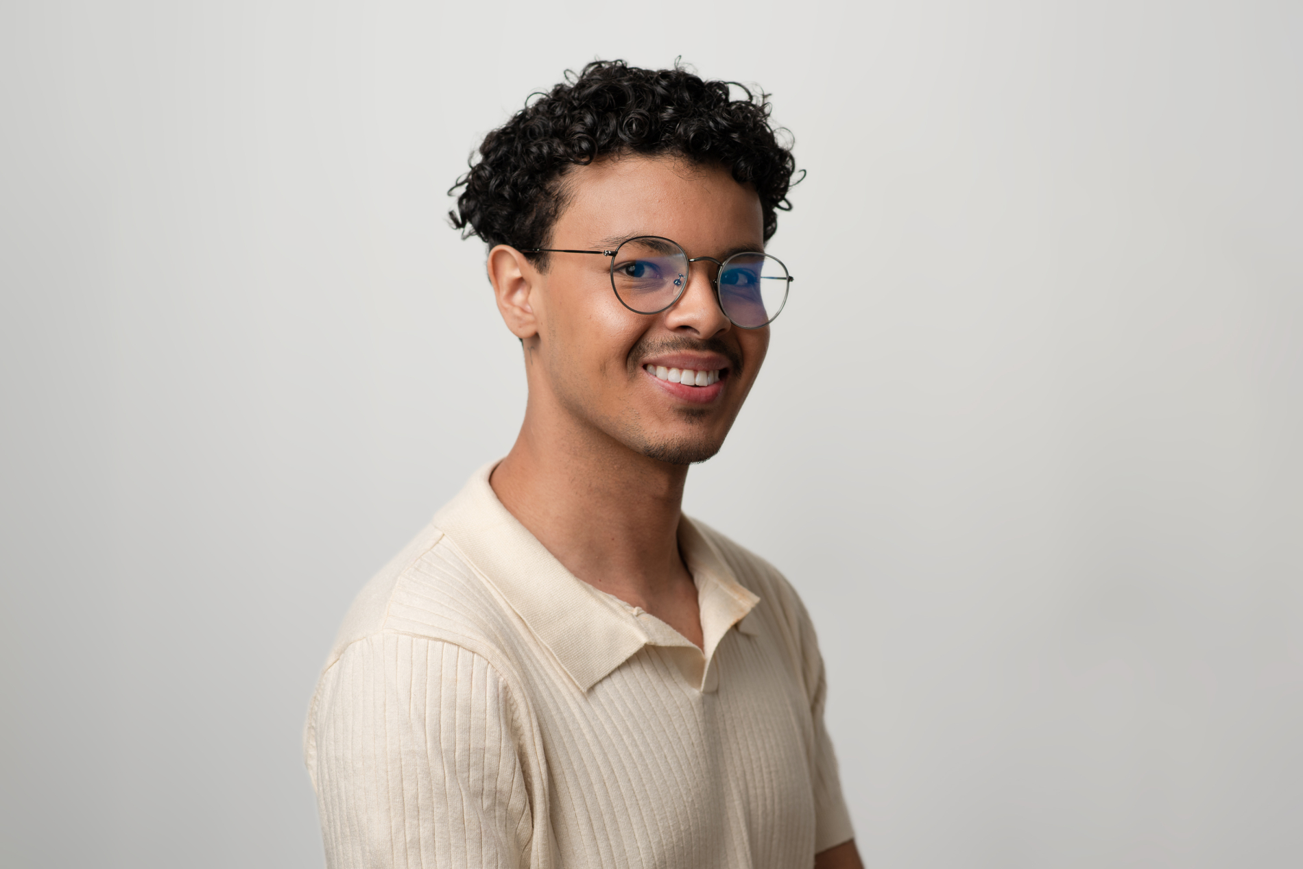 A man smiling widely. He has dark, curly hair, round glasses, and wears an off-white collared shirt.