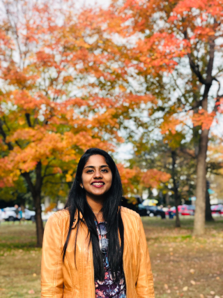Young woman with long black hair smiling outside surrounded by trees with red and orange leaves