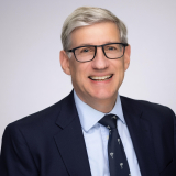 David Koppel wearing a navy blazer, light blue shirt, navy tie and glasses