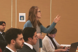 Side-view of a woman in blue top and grey dress asking a question amid an audience. She is gesturing with her hands while audience members look on.