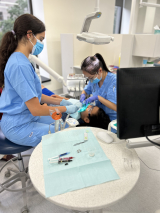 woman in blue scrubs performing dental work in a clinic on a child lying in a chair, with another woman in blue scrubs to the right providing assistance 