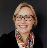 woman with blond hair tied back, wearing glasses, orange scarf, and black jacket