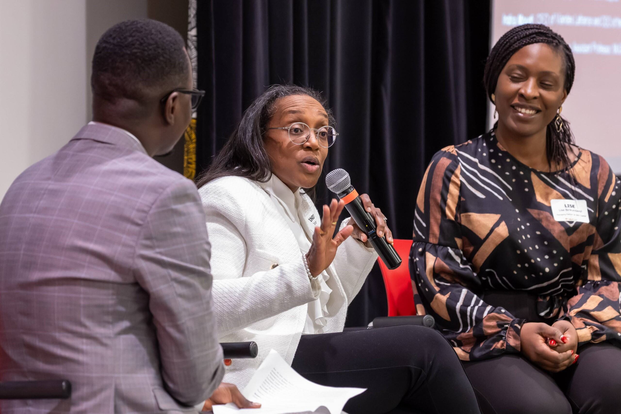 Panelist Indira Moudi speaks into microphone, in conversation with panelist Lise Birikundavyi and moderator Dr. Anicet Fangwa. 