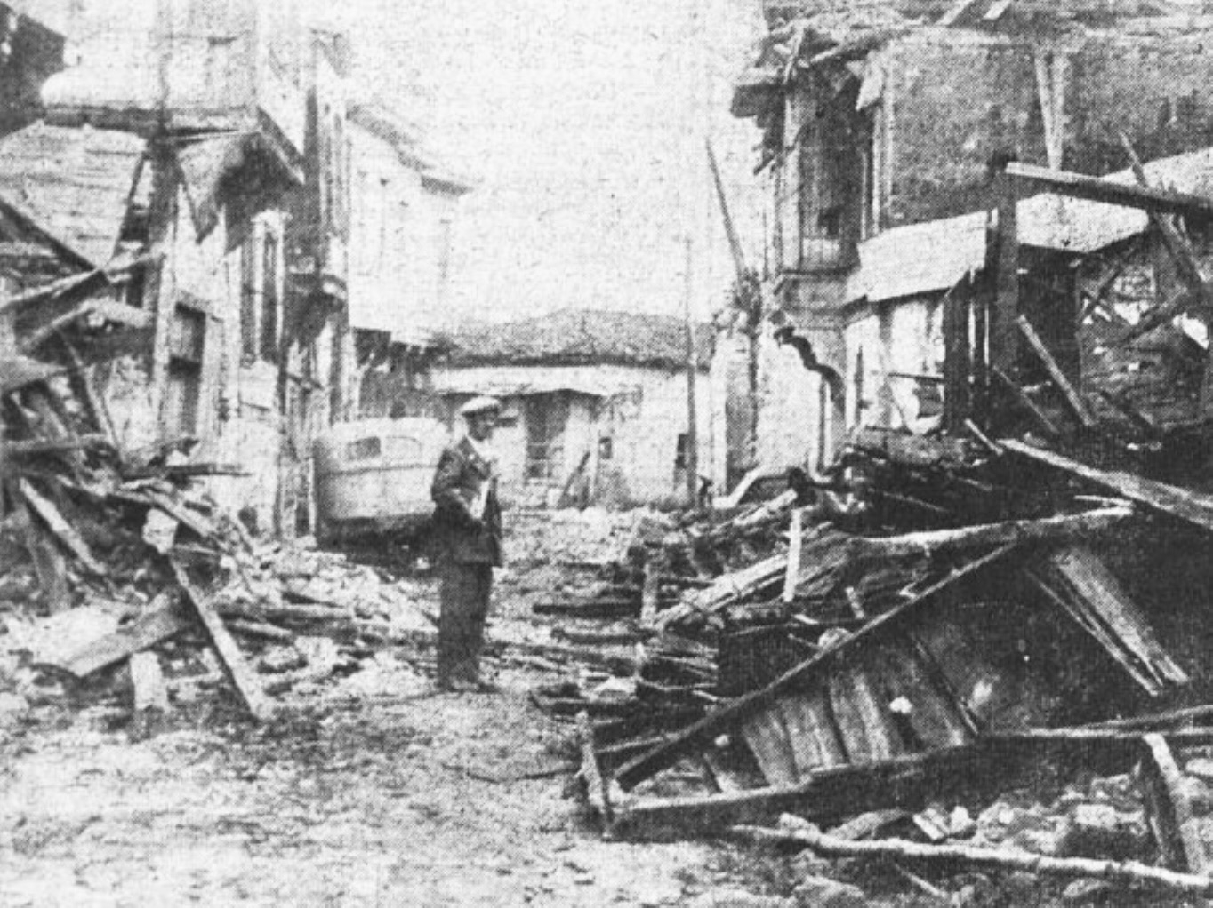 The black-and-white image shows a man standing on a road lined with collapsed buildings on both sides.