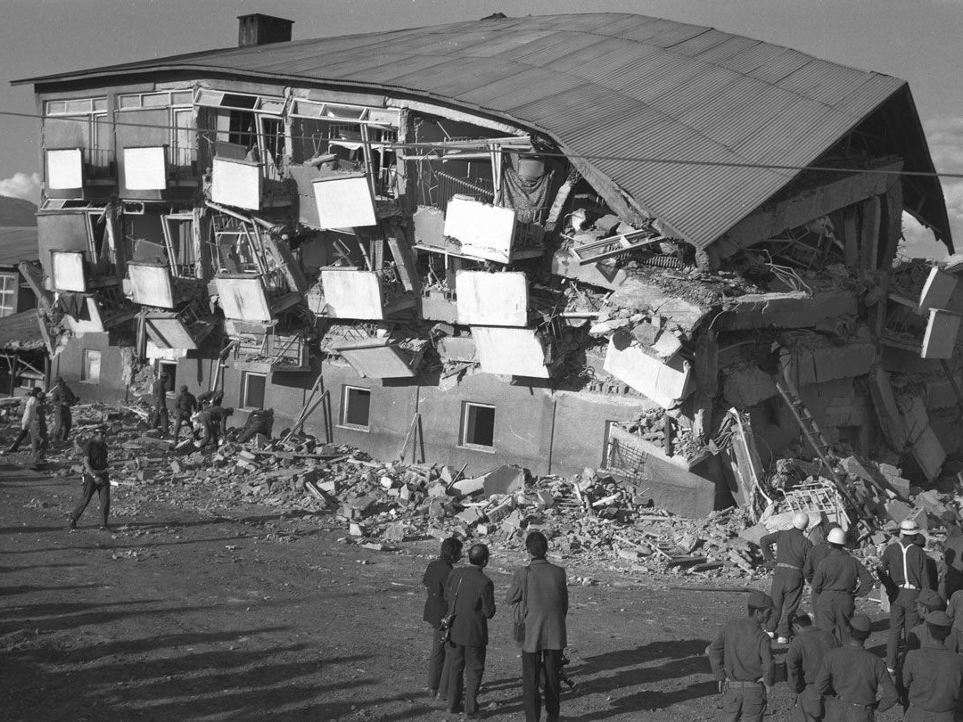 The black-and-white image shows a handful of people walking on the street along a ruined site with a collapsed building.