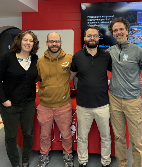 Four people standing together in front of a red wall and presentation screen.