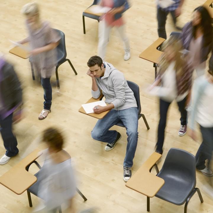 Troubled male university student sits at a desk while the rest of the class appears blurry