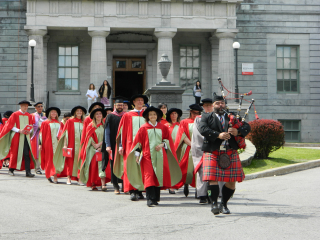 Group of McGill faculty in red robes walking behind man playing the bagpipe in front of school building