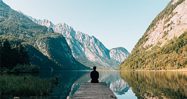 A person sitting on a dock near a lake, surrounded by mountains