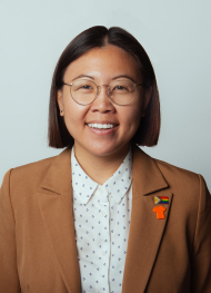 Headshot of Michelle Lui smiling directly at camera wearing a polka dot shirt and brown blazer with New Progress Pride Flag pin and an Orange Shirt pin on a white backdrop