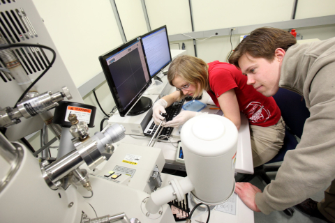2 students looking into a machine in a lab, with a desktop in front