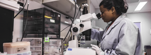 A student wearing a lab coat looking into a microscope in a lab