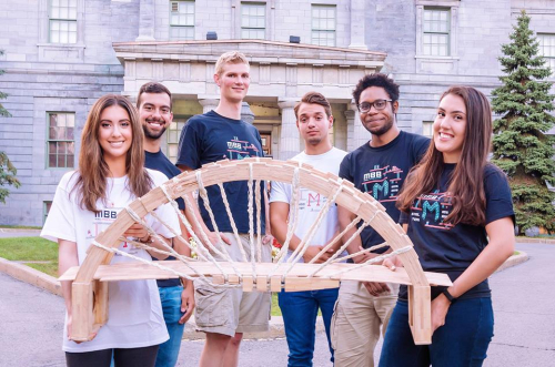 A group of students holding a small bridge made out of wood in front of Arts building in downtown campus