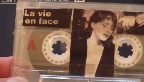 A hand is holding an audio cassette with the writing "La vie en face" and a black and white image of a woman.