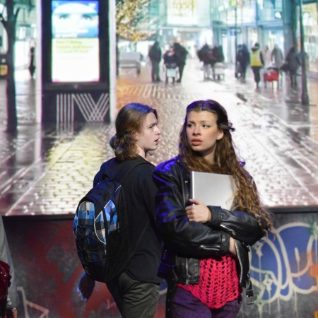 Two young people with black jackets and a young person impersonating an older woman look concerned. In the back we see a video projection of downtown Montreal at night.