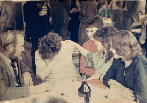 four woman and a man sitting at a table, one is distressed and the rest are comforting her.