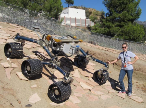 A smiling woman wearing sunglasses with a grey t-shirt, jeans with a lanyard around her neck places her hand on a 6-wheeled rover on a slope 