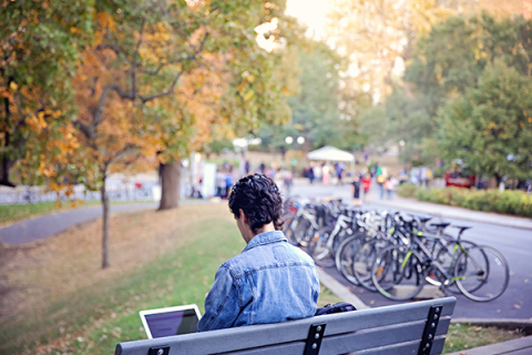Young man sitting on a bench with his laptop in a campus setting