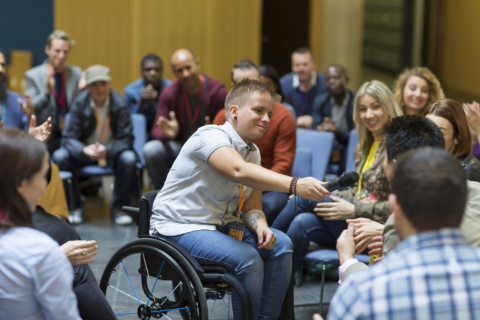 Un étudiant en fauteuil roulant tendant un microphone à un autre participant pendant une assemblée publique.