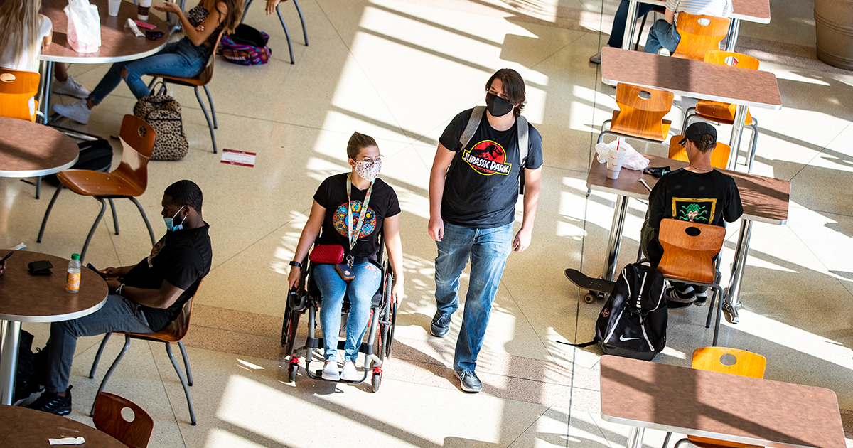 Students in a large sunlit space. Two students, one in a wheelchair, pass by occupied tables.