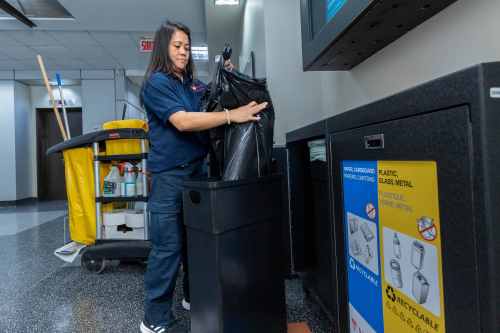 A woman takes out a garbage bag of a black bin standing in front of a multi-stream sorting station.