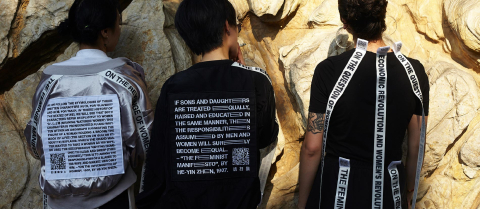 3 people standing against a wall, facing away from the camera wearing clothing with small printed text.