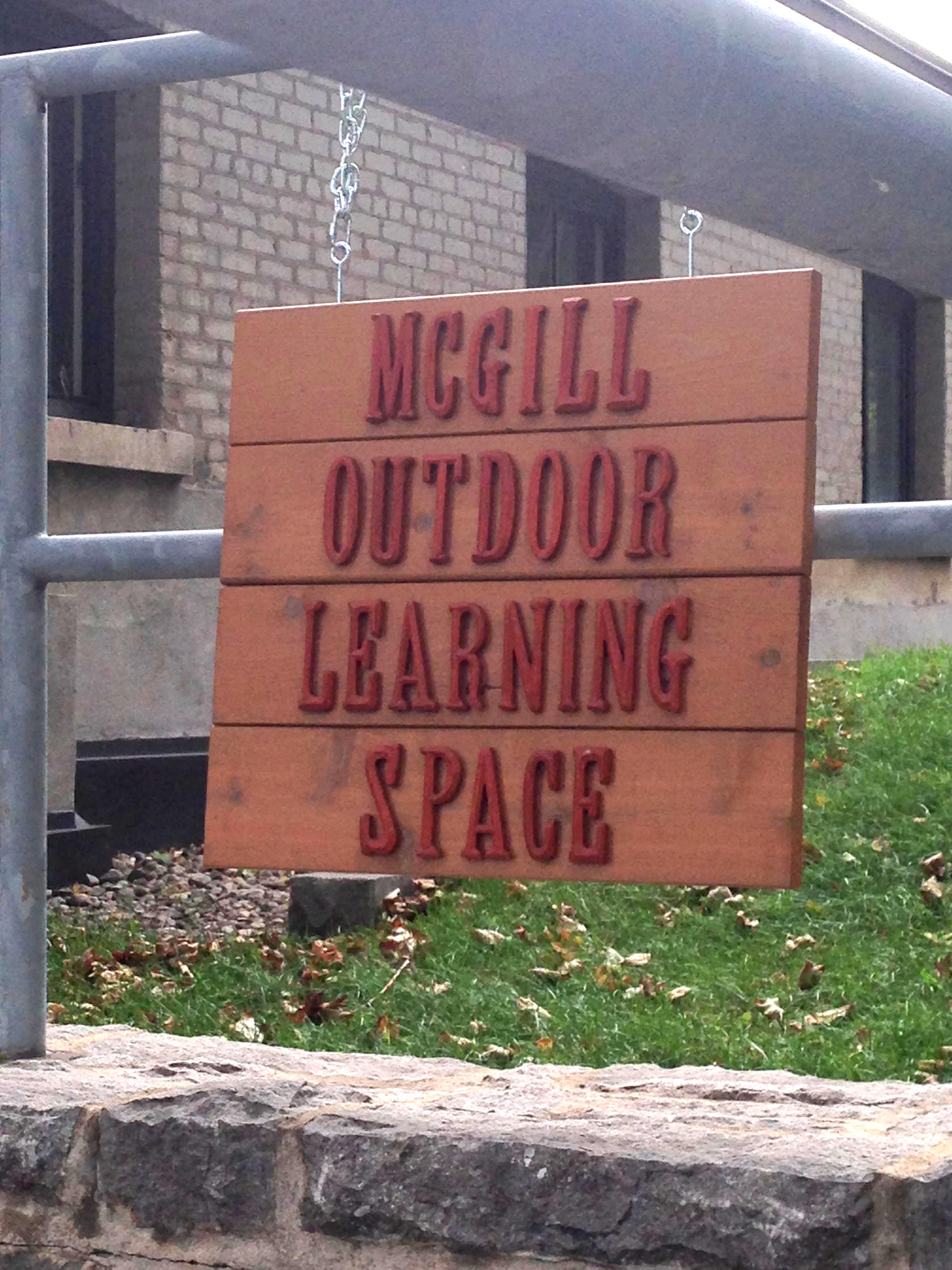 A wooden sign that reads McGill Outdoor Learning Space in raised letters hangs above grass and a brown building.