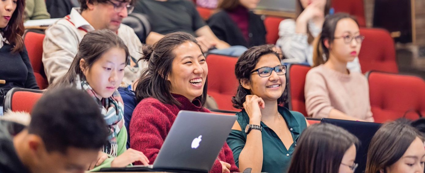 three smiling students sit in a large classroom, one looking at their laptop and the others looking towards their professor