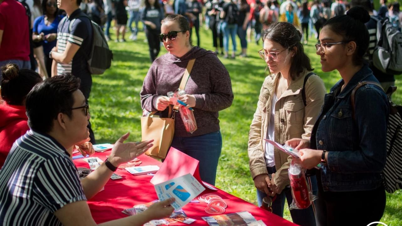 three students stand outdoors at a table speaking to a staff member about activities on campus