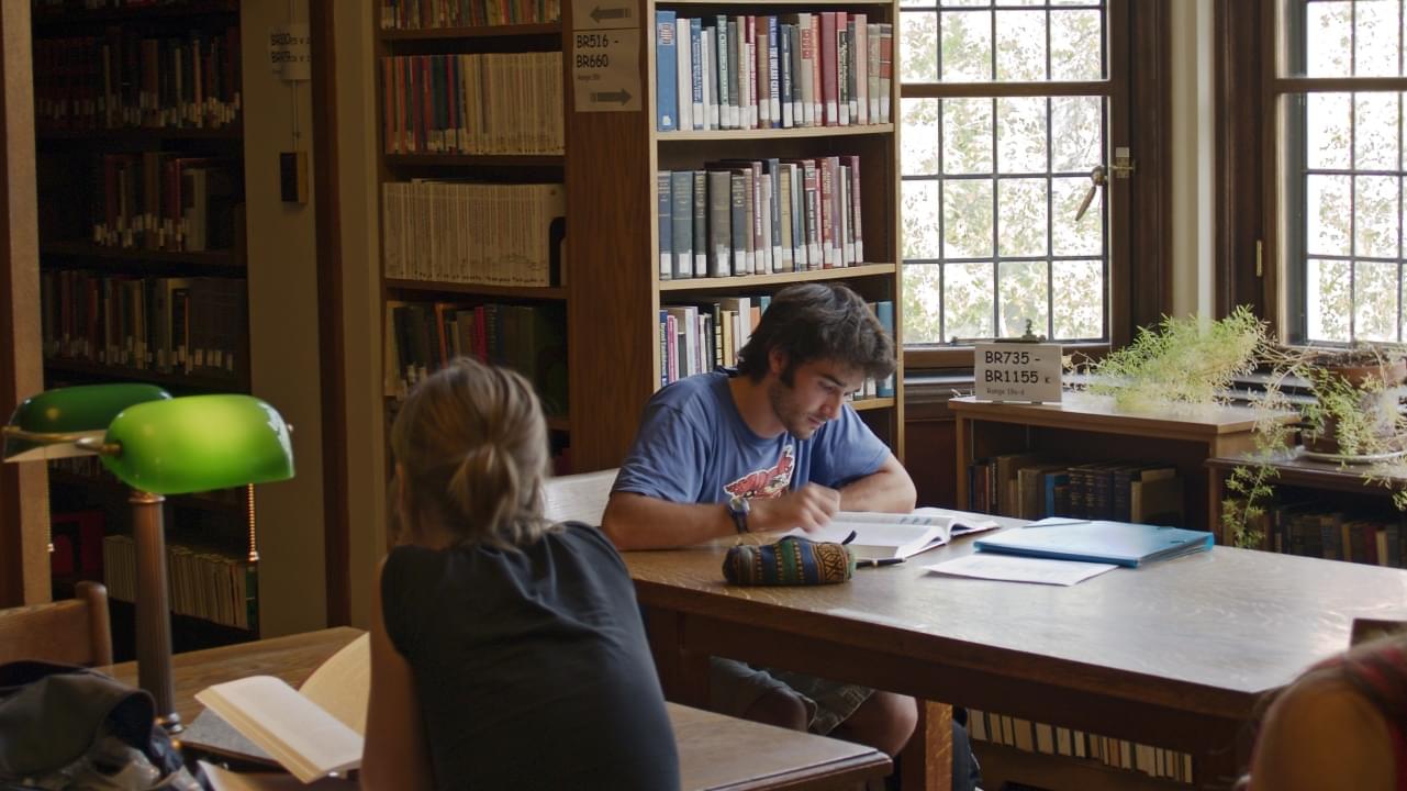 a student is seated in a library near a window reading a book