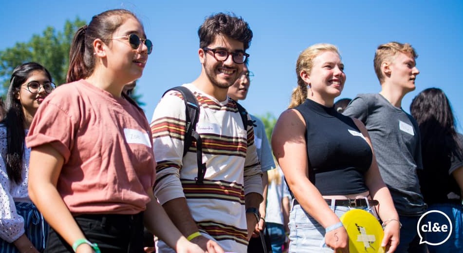 a group of students stand outside on a sunny day listening to a speaker who is out of view